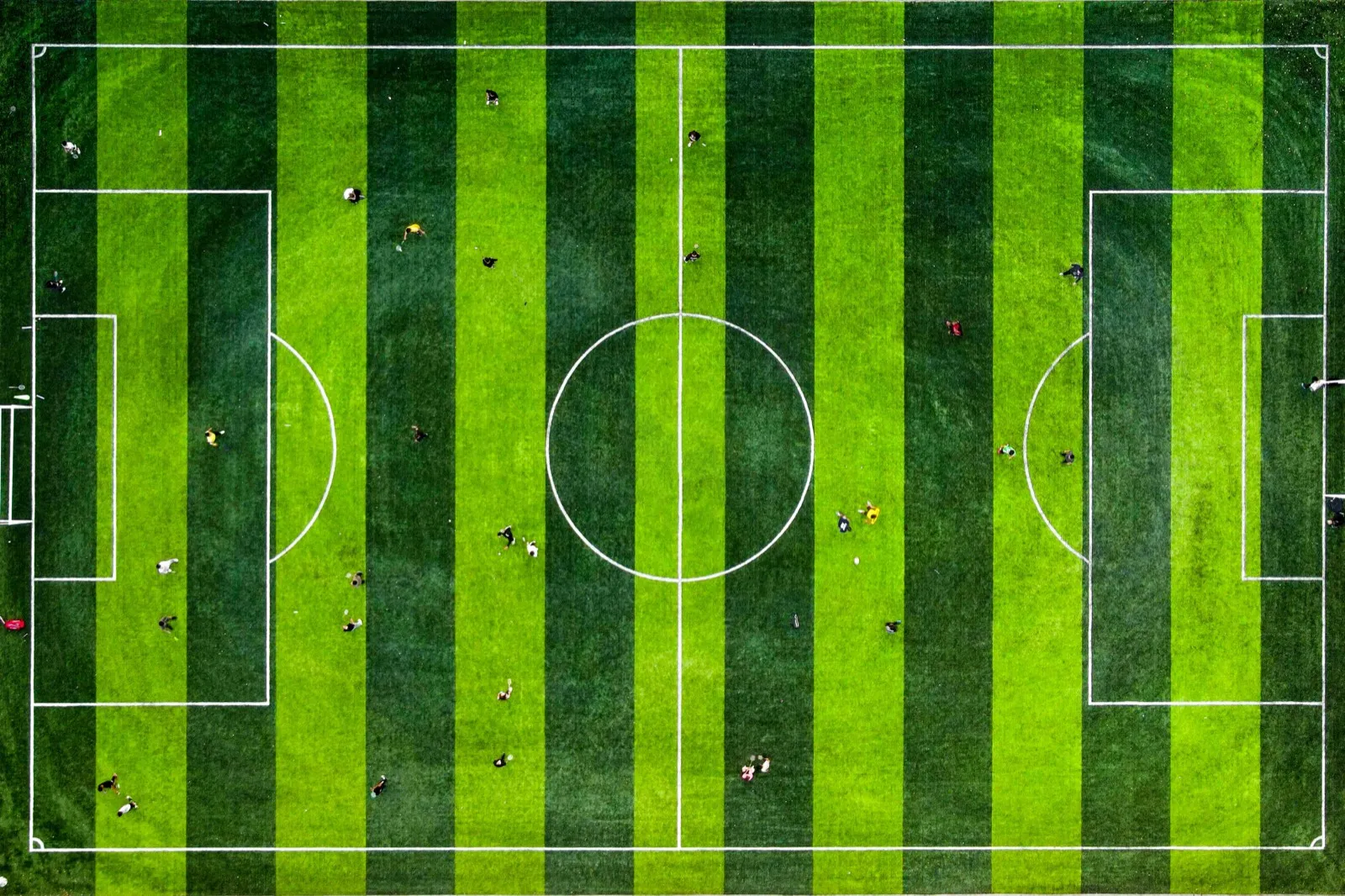 Luftaufnahme eines Fußballplatzes mit Mittelkreis und Strafräumen, auf dem einzelne Spieler stehen.